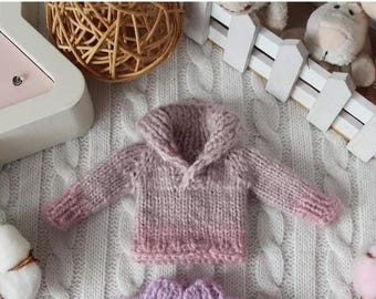 May include: A small, hand-knitted pink and lavender sweater with a hood. The sweater is made of soft yarn and has a gradient color effect. The sweater is displayed on a white cable-knit blanket, with decorative items in the background.