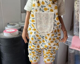 May include: A doll wearing a white shirt with a lemon print overall. The overall has a large pocket on the front. The doll is wearing white socks and sneakers.