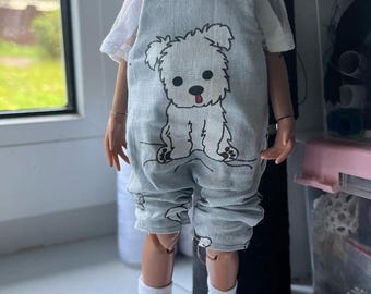 May include: A doll wearing a light blue denim overall with a white t-shirt and a white dog graphic. The doll is wearing white socks and pink and black sneakers.