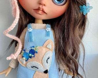 May include: A Blythe doll with large blue eyes and long brown hair. The doll wears a light blue overall dress with a felt fox applique and a pink knit hat. A blue flower hair clip adds a decorative touch. The doll's face is painted with rosy cheeks and a neutral expression.