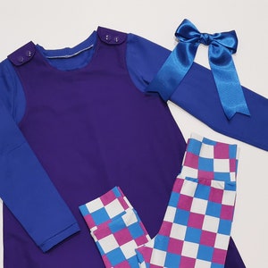 Puede incluir: Un vestido largo de manga larga morado con una camisa de manga larga azul debajo. El vestido tiene dos botones en los hombros. Un lazo azul se encuentra en el lado derecho de la imagen. Unos leggings a cuadros rosa y azul se encuentran en la parte inferior de la imagen.