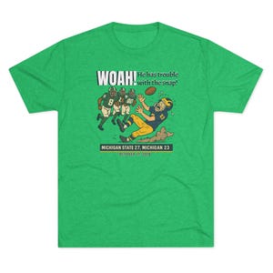 May include: A green t-shirt featuring a cartoon illustration of a football play. The graphic includes the text "WOAH! He has trouble with the snap!" and the score of a game: MICHIGAN STATE 27, MICHIGAN 23, October 17, 2015.