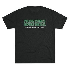 May include: Dark gray t-shirt with the text "PRIDE COMES BEFORE THE FALL" in green, attributed to Mark Dantonio, 2007. The shirt is a crew neck style and appears to be made of a soft, comfortable material.
