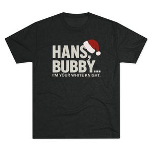 May include: Dark grey t-shirt featuring the text "HANS, BUBBY... I'M YOUR WHITE KNIGHT." in white, with a Santa hat graphic. The shirt has a crew neck and short sleeves.