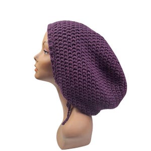 May include: A dark purple crochet hat with a textured pattern, displayed on a mannequin head. The hat features a drawstring and is handmade. The hat's design is visible. The hat is a deep shade of purple.