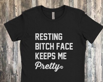 May include: Black t-shirt with the white text "RESTING BITCH FACE KEEPS ME Pretty". The shirt is a crew neck style and appears to be made of a soft material. The text is centered on the front of the shirt.