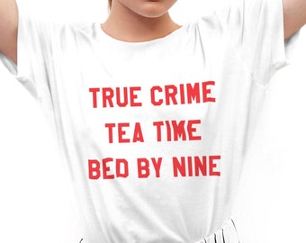 Op de afbeelding: Wit T-shirt met rode tekst die "True Crime Tea Time Bed By Nine" luidt. De persoon draagt een witte broek met strepen.