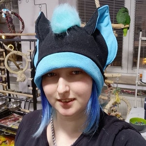 May include: A person with blue hair is wearing a black and blue fleece hat with fuzzy blue ear flaps and a blue pom-pom on top.