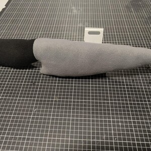 May include: A soft toy knife featuring a black handle and a grey blade. The toy, made from soft fabric, is designed to mimic a real knife. It rests on a black and white grid surface.
