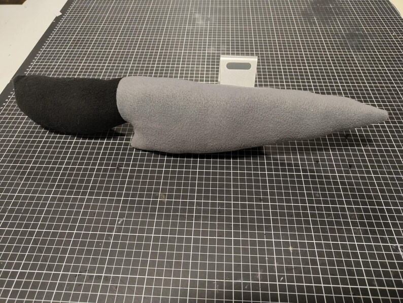 May include: A soft toy knife featuring a black handle and a grey blade. The toy, made from soft fabric, is designed to mimic a real knife. It rests on a black and white grid surface.