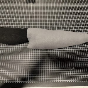 May include: A soft toy knife featuring a black handle and a grey blade. The toy is constructed from soft fabric and mimics the appearance of a real knife. It rests on a black cutting mat.