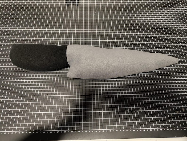 May include: A soft toy knife featuring a black handle and a grey blade. The toy is constructed from soft fabric and mimics the appearance of a real knife. It rests on a black cutting mat.