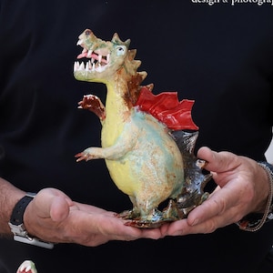 Pu&ograve; includere: Statuine di draghi in ceramica fatte a mano. I draghi sono dipinti in giallo, verde e marrone, con accenti rossi a forma di ali. I draghi a bocca aperta hanno denti visibili. Le statuine sono esposte su una superficie bianca.