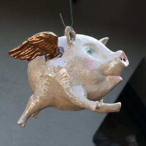 Könnte beinhalten: Eine skurrile Schweinefigur aus Keramik mit goldenen Flügeln, die an einem Faden hängt. Das Schwein hat einen hellrosa Körper, blaue Augen und eine detaillierte Schnauze. Die Flügel sind goldfarben, was den Eindruck von Flug erweckt.