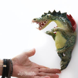 Pu&ograve; includere: Una scultura in ceramica di un alligatore con corpo verde e giallo, dettagli rossi e denti affilati. La scultura &egrave; montata su un muro bianco. L'alligatore sembra interagire con una mano.