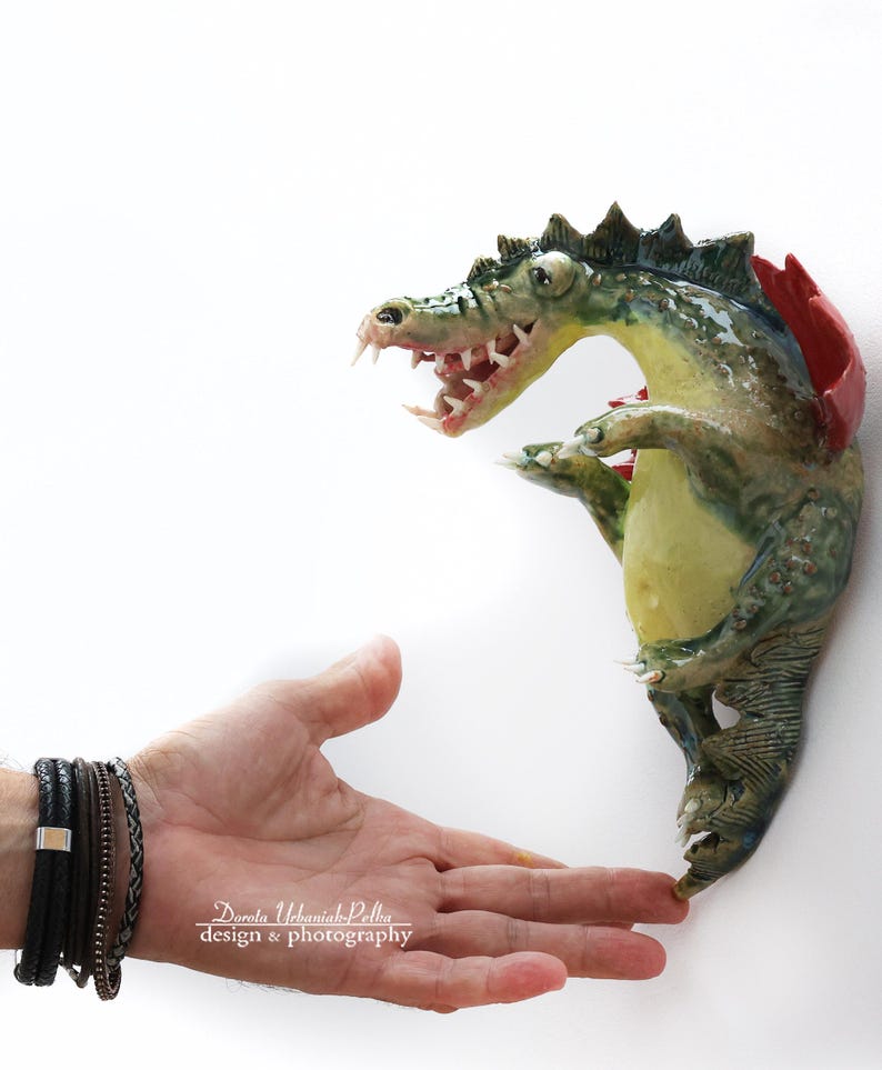 Pu&ograve; includere: Una scultura in ceramica di un alligatore con corpo verde e giallo, dettagli rossi e denti affilati. La scultura &egrave; montata su un muro bianco. L'alligatore sembra interagire con una mano.
