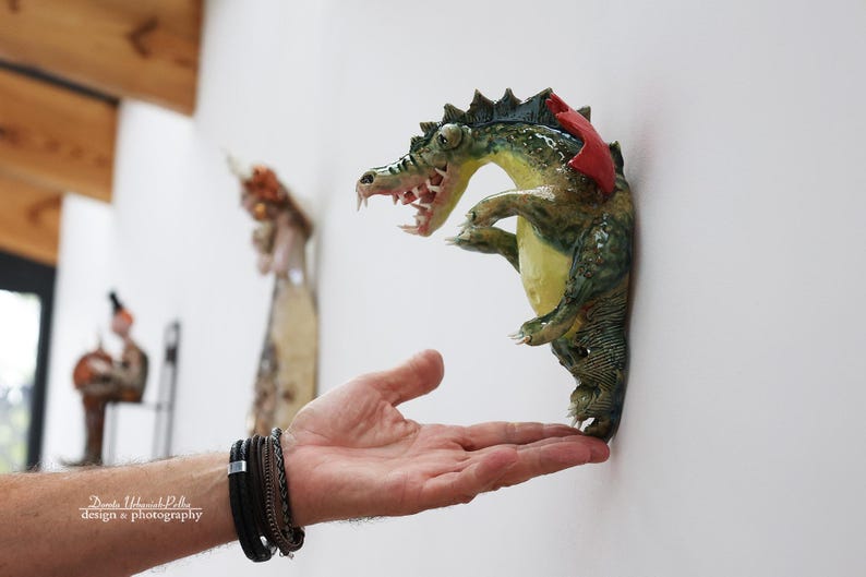 Pu&ograve; includere: Scultura da parete in ceramica a forma di alligatore verde, giallo e rosso. L'alligatore ha la bocca aperta con denti visibili ed &egrave; montato su una parete bianca. Fa parte di una collezione d'arte.