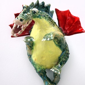 Pu&ograve; includere: Scultura di drago in ceramica con pancia gialla, squame verdi e blu e ali rosse. Il drago ha la bocca aperta con denti bianchi ed &egrave; posizionato come se fosse in piedi. La scultura &egrave; montata su una superficie bianca.