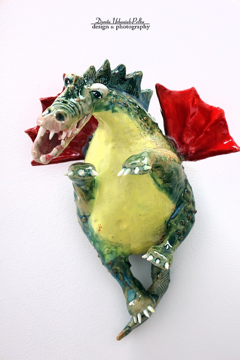 Pu&ograve; includere: Scultura di drago in ceramica con pancia gialla, squame verdi e blu e ali rosse. Il drago ha la bocca aperta con denti bianchi ed &egrave; posizionato come se fosse in piedi. La scultura &egrave; montata su una superficie bianca.