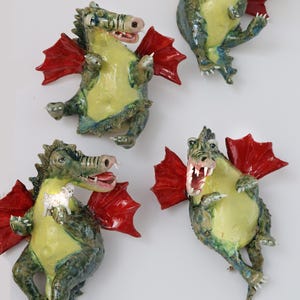 Pu&ograve; includere: Quattro figurine di draghi in ceramica con ali rosse, pance gialle e dettagli verdi e blu. Un drago tiene in mano un piccolo agnello bianco. I draghi hanno la bocca aperta e sono in varie pose.