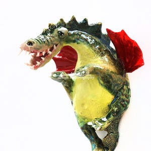 Pu&ograve; includere: Scultura di drago in ceramica fatta a mano nei toni del verde, giallo e blu, con un'ala rossa. Il drago ha la bocca aperta con denti visibili. La scultura &egrave; firmata "Dorota Urbaniak-Pelka design & photography".
