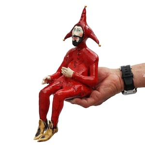 May include: A ceramic figurine of a jester wearing a bright red suit and a matching three-pointed hat with small gold-colored tips. The figure has a painted face with a dark beard, light-colored hands, and pointed tan shoes, depicted in a seated pose.