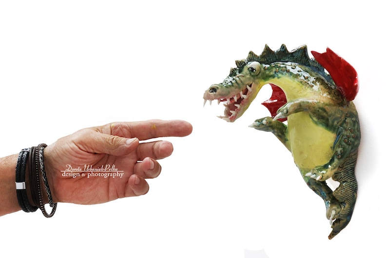 Pu&ograve; includere: Una stravagante scultura di alligatore dipinta a mano con un'ala rossa, corpo verde e giallo e bocca aperta. L'alligatore sembra emergere da un muro. Una mano con braccialetti punta verso l'alligatore.