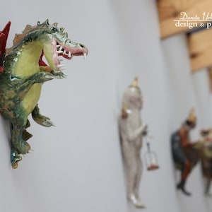 Pu&ograve; includere: Scultura di drago in ceramica montata a parete con un'ala rossa e bocca aperta, che mostra tonalit&agrave; verdi, gialle e marroni. Altre statuette sono visibili sullo sfondo. L'immagine &egrave; di Dorota Urbaniak-Pelka design & photography.