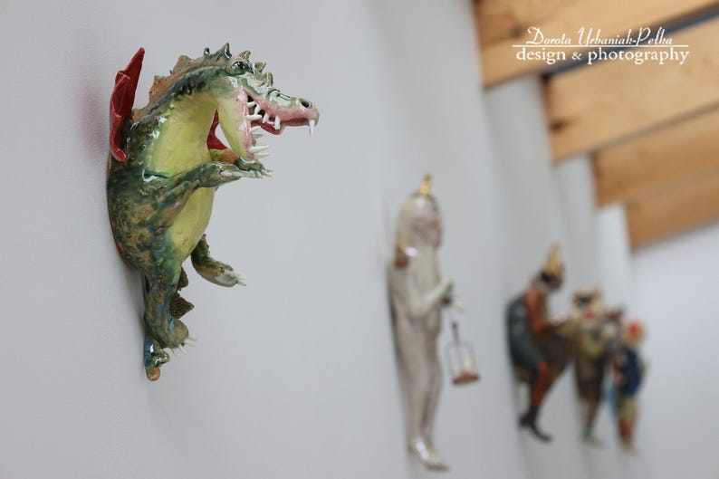Pu&ograve; includere: Scultura di drago in ceramica montata a parete con un'ala rossa e bocca aperta, che mostra tonalit&agrave; verdi, gialle e marroni. Altre statuette sono visibili sullo sfondo. L'immagine &egrave; di Dorota Urbaniak-Pelka design & photography.