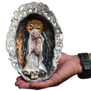 Może przedstawiać: Ceramiczna tablica ścienna przedstawiająca wyrzeźbioną postać trzymającą dziecko, umieszczoną w ciemnej, opalizującej owalnej misie. Całość otoczona jest misterną, białą obwódką przypominającą pnącza i posiada złocistą aureolę za postacią.