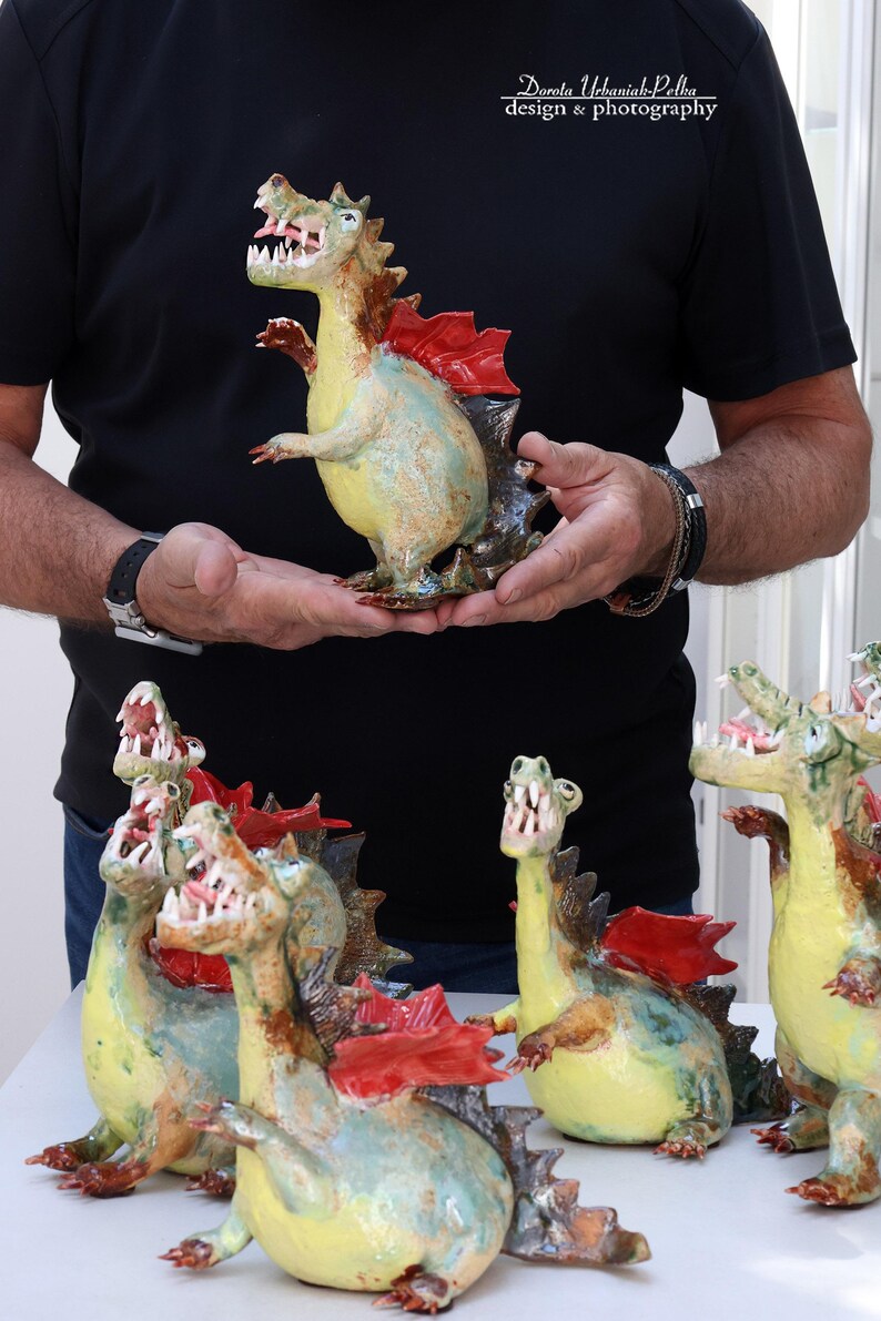 Pu&ograve; includere: Statuine di draghi in ceramica fatte a mano. I draghi sono dipinti in giallo, verde e marrone, con accenti rossi a forma di ali. I draghi a bocca aperta hanno denti visibili. Le statuine sono esposte su una superficie bianca.