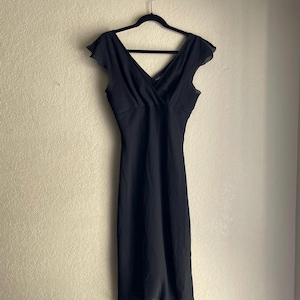 May include: A black midi dress with a V-neckline and cap sleeves, hanging on a black hanger against a neutral wall. The dress has a flowing skirt and a fitted bodice, suitable for formal occasions.