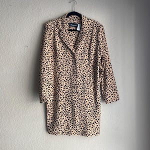 May include: A tan blazer with a black animal print pattern hangs on a black hanger. The blazer has a collared neckline, three-quarter sleeves, and a button-down front. The garment is made of a lightweight fabric.