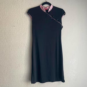 May include: Black dress with a mandarin collar and cap sleeves. The dress features a pink satin trim along the collar, sleeves, and a decorative button closure. The dress is hanging on a black hanger against a neutral wall.