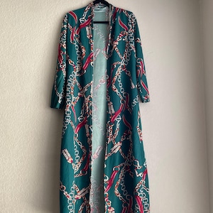 May include: A long, open-front duster in a teal green color, adorned with a repeating pattern of gold chains and red accents. The duster is hanging on a black hanger against a neutral wall.