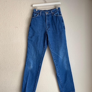 Op de afbeelding: Blauwe spijkerbroek hangend aan een witte hanger. De jeans heeft een klassiek ontwerp met vijf zakken, een knoop- en ritssluiting en een rechte pijp. De denim heeft wat vervaging en slijtage.