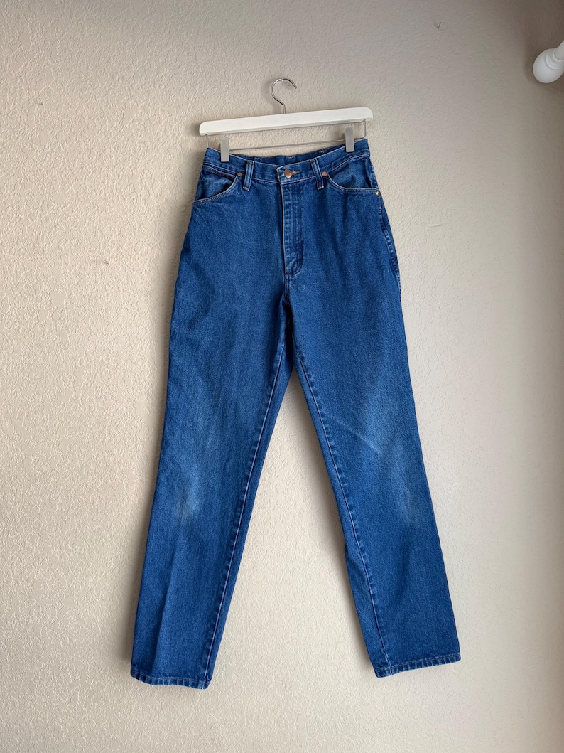 Op de afbeelding: Blauwe spijkerbroek hangend aan een witte hanger. De jeans heeft een klassiek ontwerp met vijf zakken, een knoop- en ritssluiting en een rechte pijp. De denim heeft wat vervaging en slijtage.