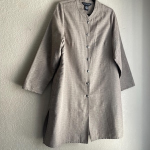 May include: A long-sleeved, button-down, beige tunic with a mandarin collar. The tunic is hanging on a black hanger against a neutral wall. The brand tag reads "Dialogue". The tunic has a textured appearance.