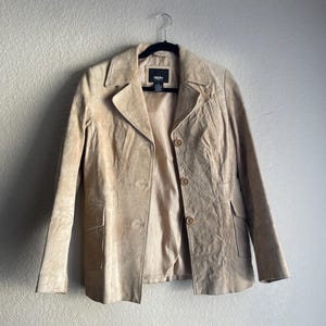 May include: A tan suede jacket with a collared neckline, button closure, and two front pockets. The jacket is hanging on a black hanger against a neutral wall. The jacket has a tailored fit.