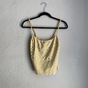 May include: A light yellow camisole top with thin straps, hanging on a black hanger. The top features a pattern of small, evenly spaced gold-colored circular cutouts across the front.
