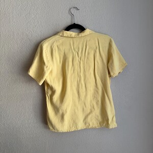 May include: A short-sleeved, pale yellow shirt with a collar, hanging on a black hanger against a neutral wall. The shirt appears to be made of a soft fabric, with visible seams and a slightly worn look.