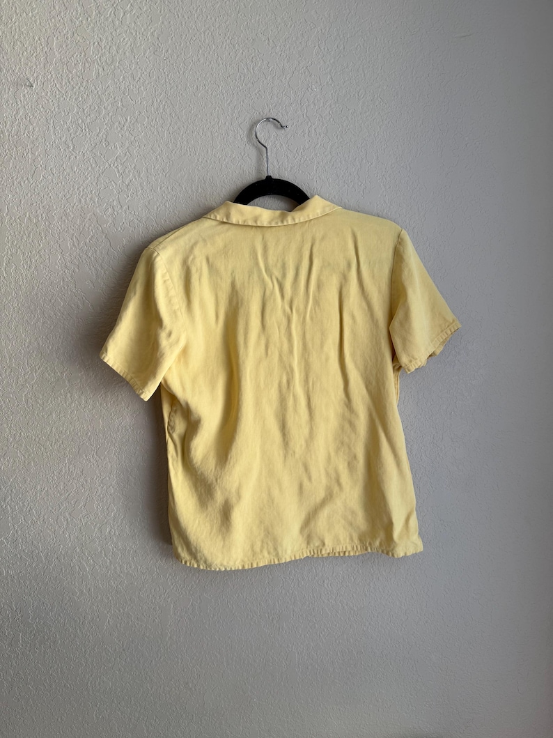 May include: A short-sleeved, pale yellow shirt with a collar, hanging on a black hanger against a neutral wall. The shirt appears to be made of a soft fabric, with visible seams and a slightly worn look.