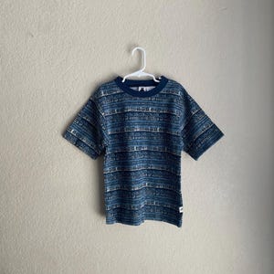May include: Short-sleeved t-shirt with a blue and white tribal pattern. The shirt has a navy blue collar and sleeves. The pattern consists of horizontal bands of geometric shapes and symbols. The shirt is hanging on a white hanger.