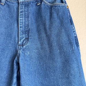 Op de afbeelding: Close-up van een blauwe spijkerbroek. De jeans heeft een knoop- en ritssluiting, koperkleurige klinknagels en een Wrangler-merklabel. De denim stof is een medium wassing, met wat slijtage en vervaging.
