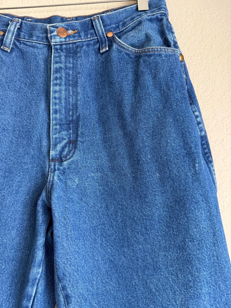 Op de afbeelding: Close-up van een blauwe spijkerbroek. De jeans heeft een knoop- en ritssluiting, koperkleurige klinknagels en een Wrangler-merklabel. De denim stof is een medium wassing, met wat slijtage en vervaging.