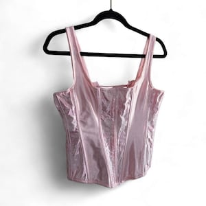 May include: A light pink corset top with a glossy, metallic finish and structured vertical paneling. The garment features wide shoulder straps, a square neckline, and a fitted bodice design, all displayed on a black plastic hanger.