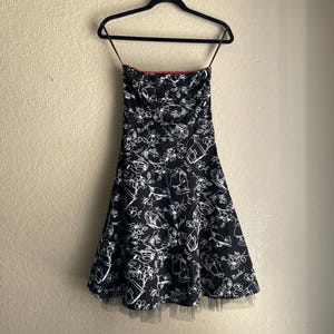 May include: A strapless black dress with a white floral and object pattern. The dress has a red trim at the top and a black tulle hem. It is hanging on a black hanger against a neutral wall.