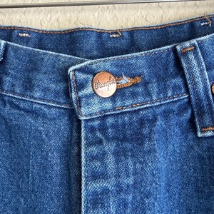 Op de afbeelding: Close-up van een blauwe denim jeans met een koperkleurige knoop. De knoop heeft het woord "Wrangler" in reli&euml;f. De jeans hangt aan een metalen hanger.