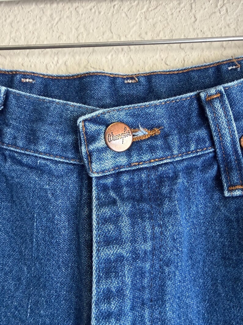 Op de afbeelding: Close-up van een blauwe denim jeans met een koperkleurige knoop. De knoop heeft het woord "Wrangler" in reli&euml;f. De jeans hangt aan een metalen hanger.