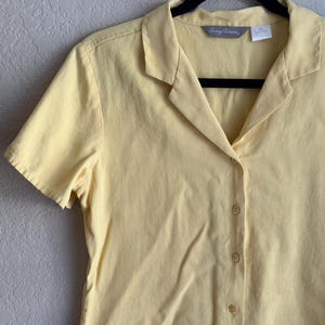 May include: A pale yellow short-sleeved button-down shirt, hanging on a black hanger. The shirt has a collared neckline and a series of small, round buttons down the front. The brand name "Tommy Bahama" is visible on the label.
